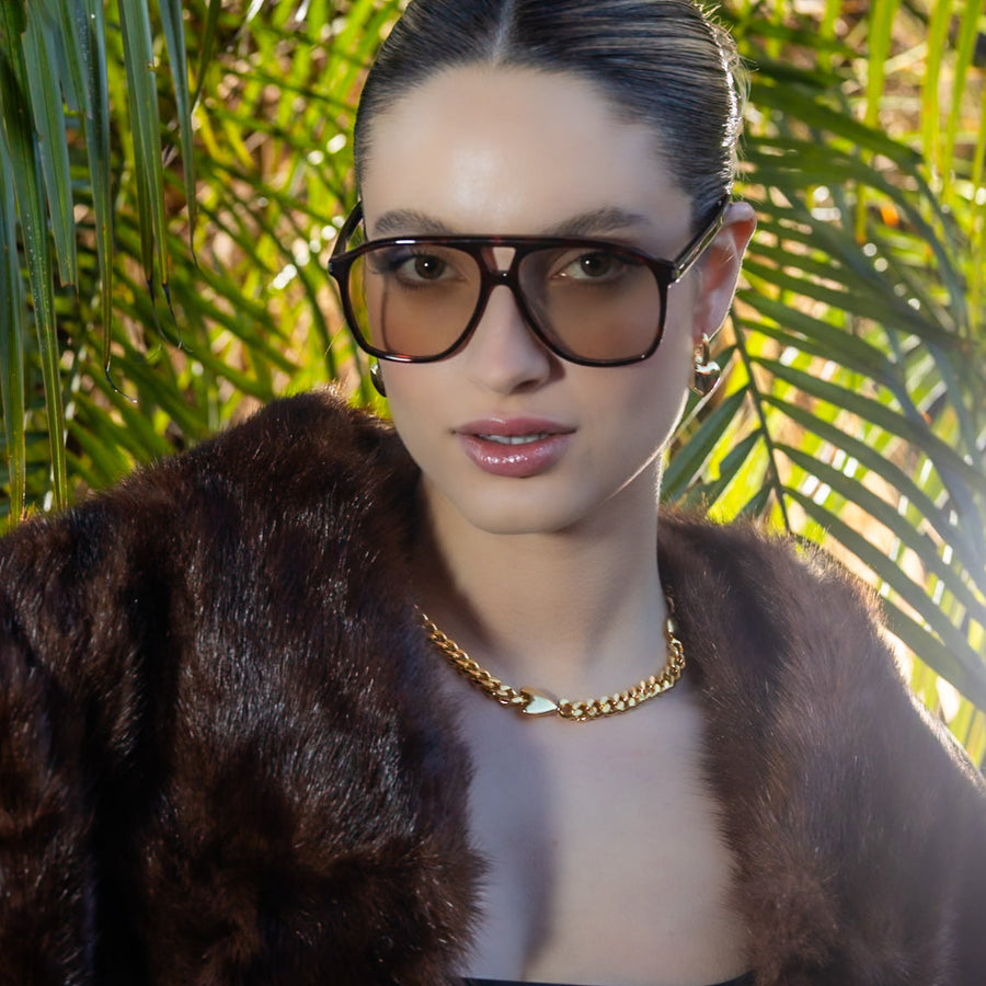 Woman wearing glasses and a fur coat with palm leaves in the background